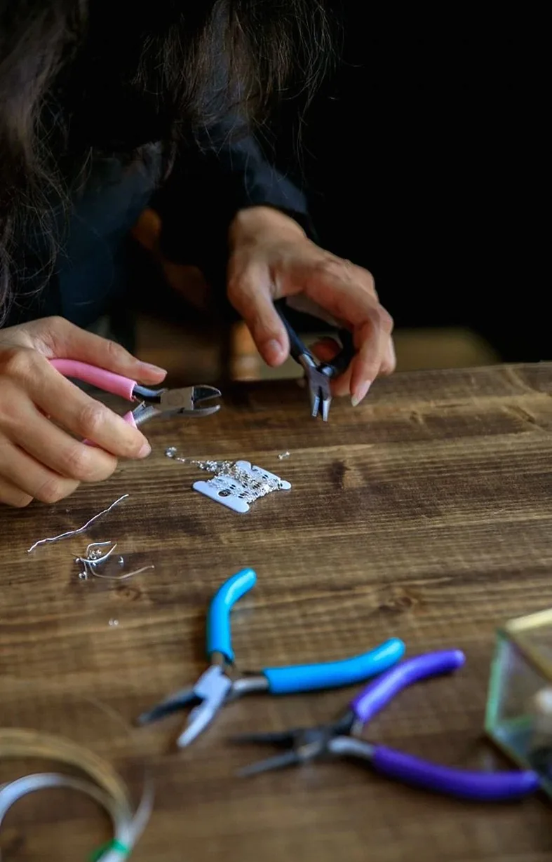 木製のテーブルでペンチとビーズを使ってジュエリーを作る女性のクローズアップ。